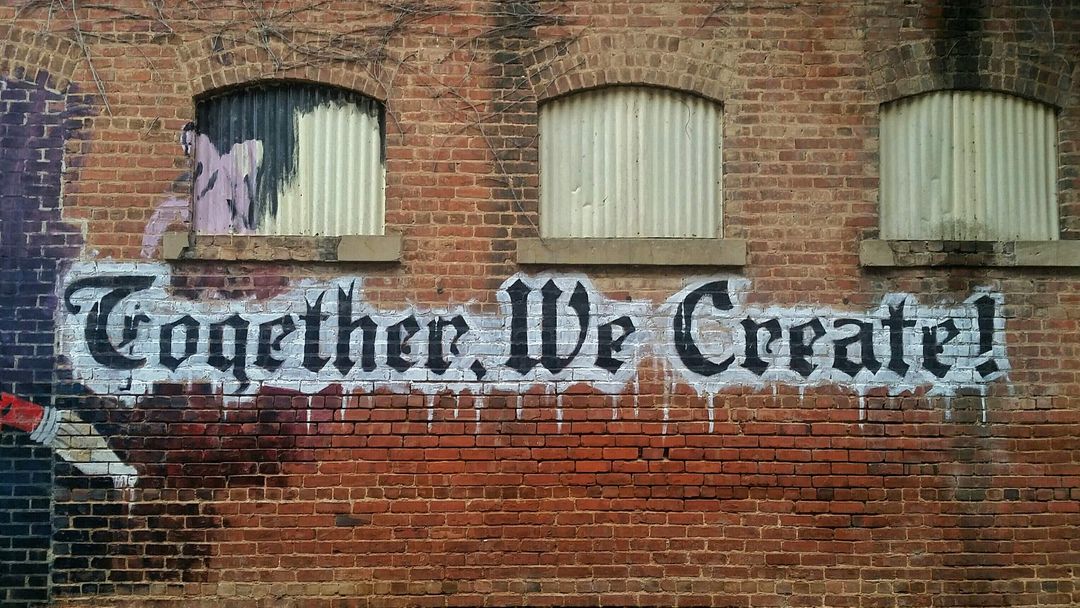 A brick building with graffiti on the side saying “Together, we create!”